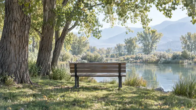 An empty wooden park bench beside a calm pond under tall cottonwood trees on a clear day, dappled sunlight on the grass and mountains in the distance: a short break waiting to be taken.